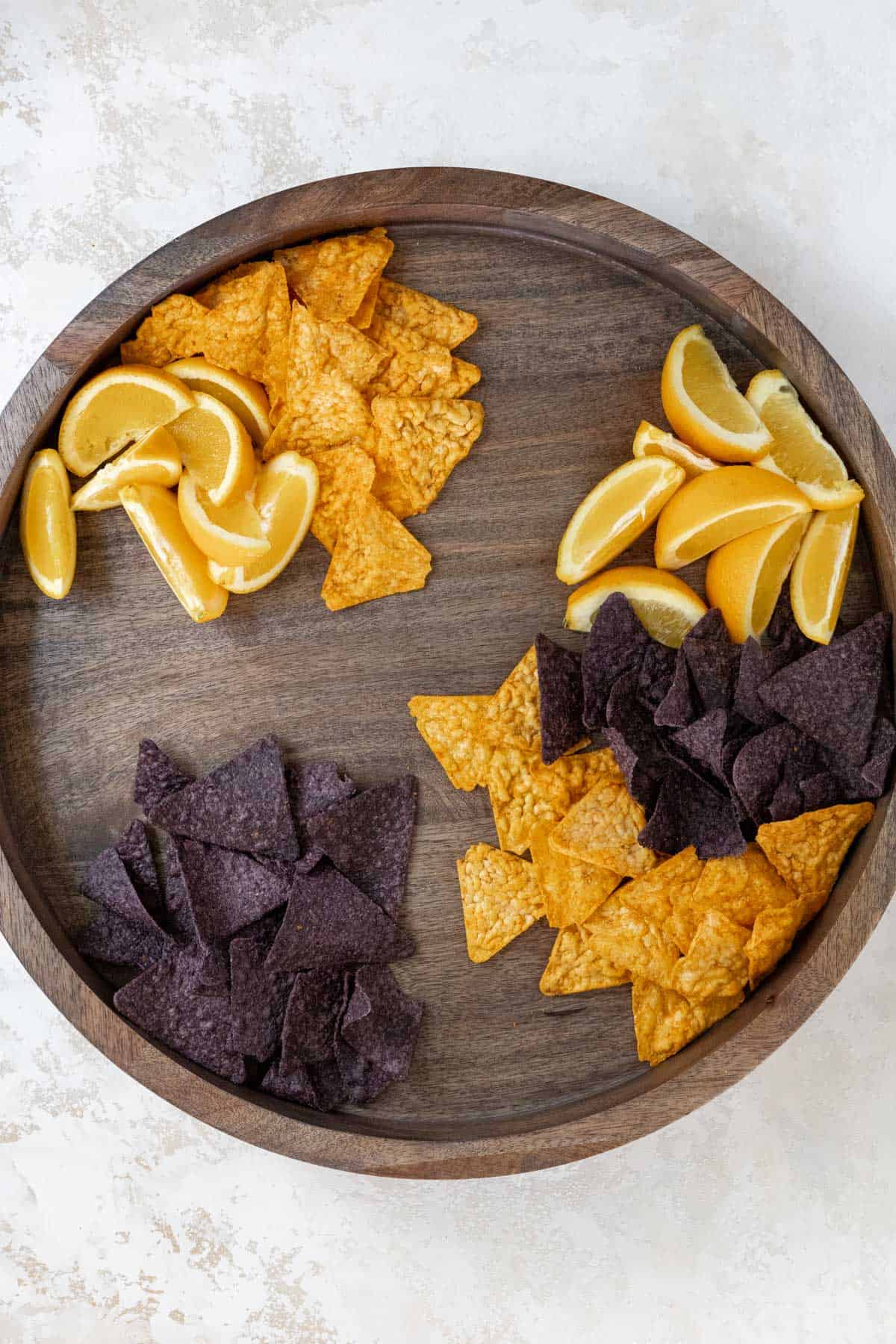 Orange wedges, blue corn tortilla chips, and nacho cheese tortilla chips arranged on a wooden board. 