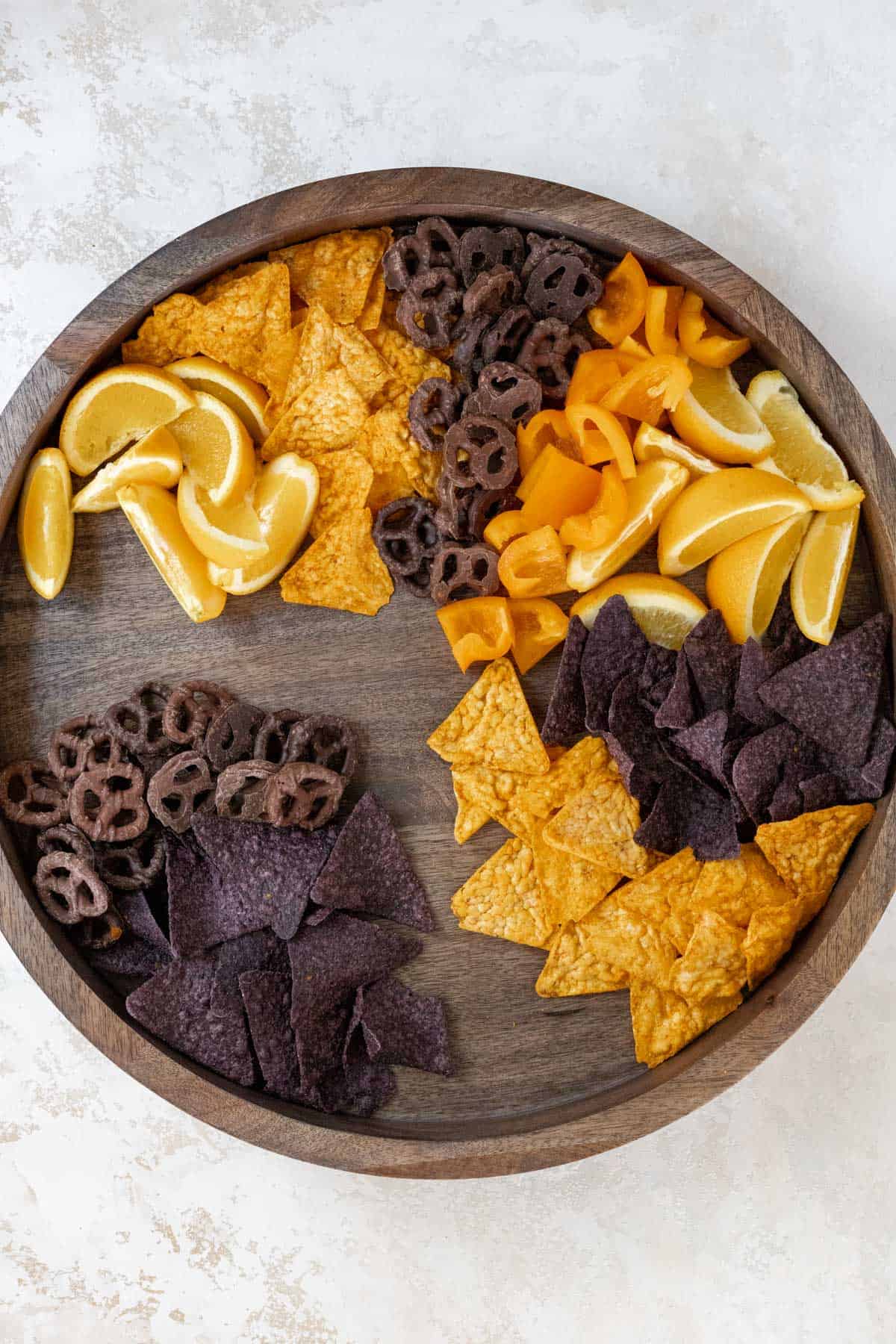 Oranges, chips, orange bell peppers, and chocolate covered pretzels arranged on a wooden board.