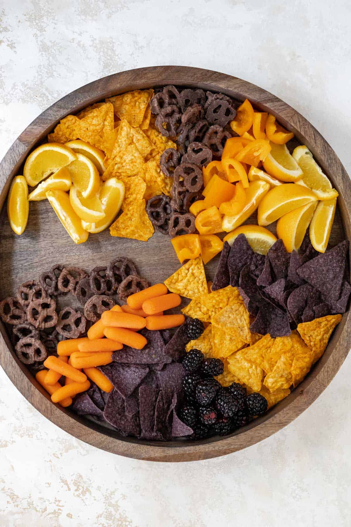 Oranges, chips, orange bell peppers, and chocolate covered pretzels, carrots, and blackberries arranged on a wooden board.