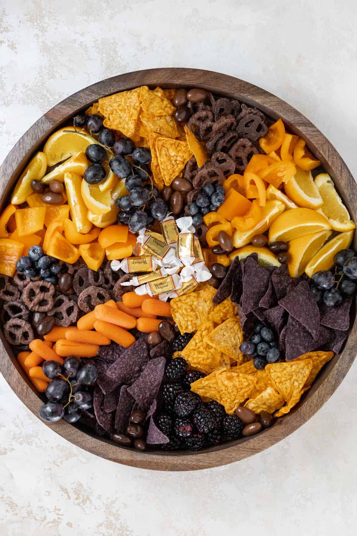 Oranges, chips, orange bell peppers, and chocolate covered pretzels, carrots, caramels, blueberries, blackberries, and chocolate almonds arranged on a wooden board.