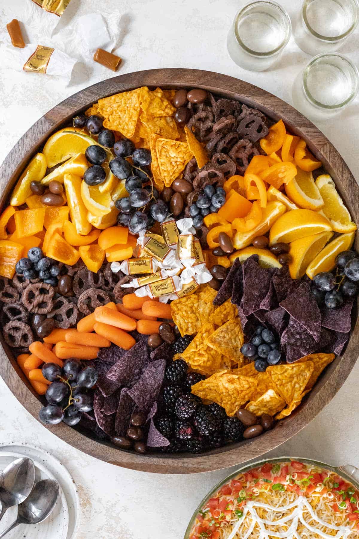 A halloween snack board on a wooden board with drinks and taco dip off to the side.