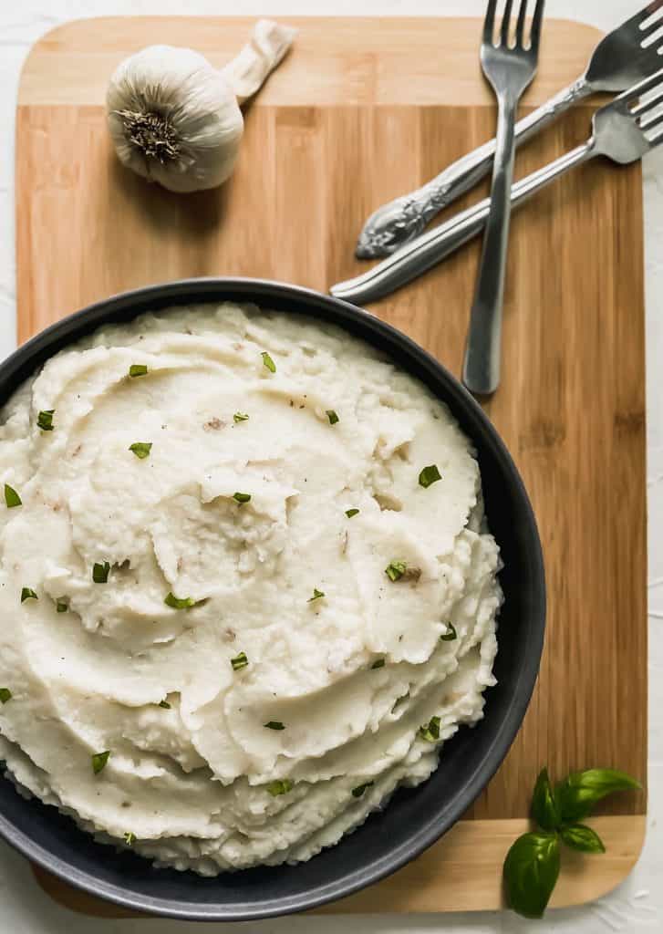 Cauliflower mash in a black bowl with a bulb of garlic and 3 forks off to the side.