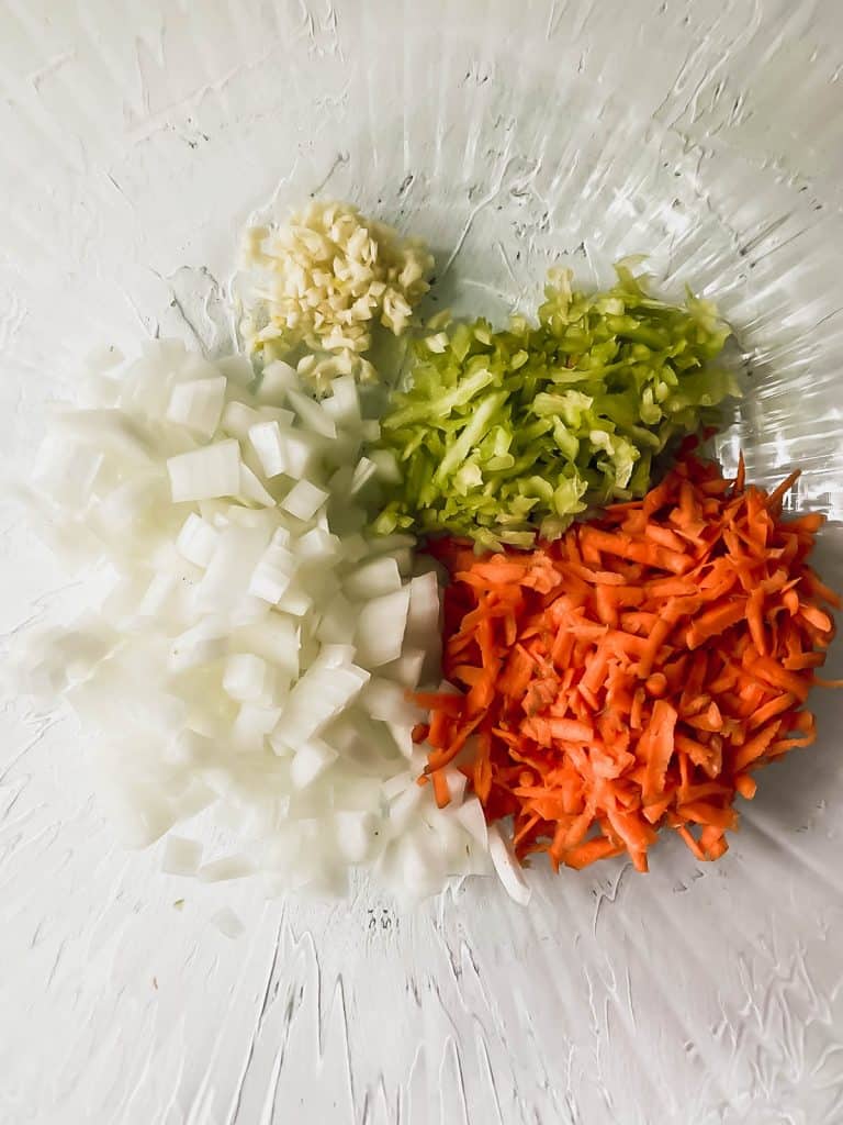 Chopped onions, minced garlic, and grated celery and carrots in a glass bowl.