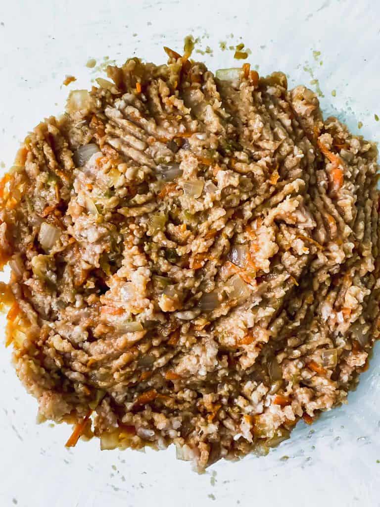 Raw ground chicken mixed with the other ingredients in a glass bowl.