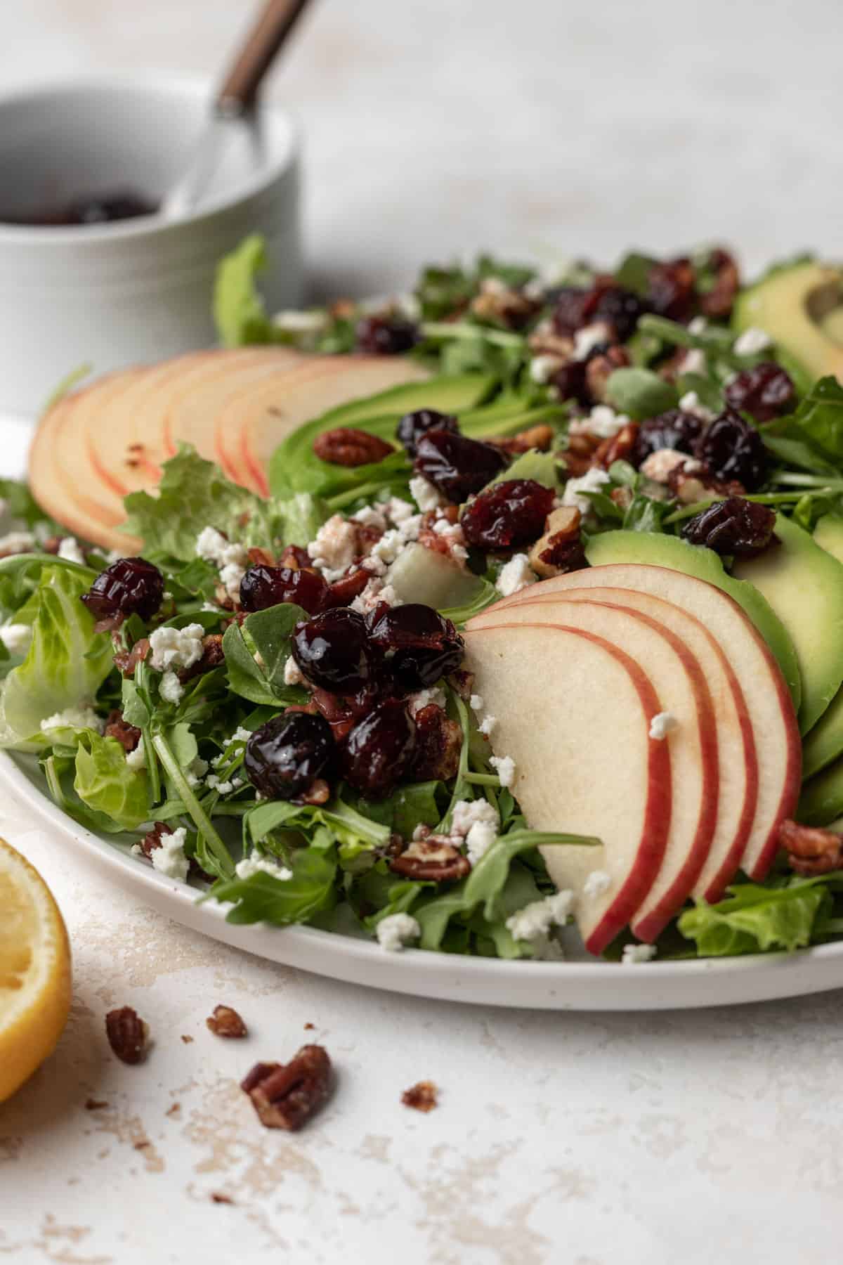 A side view of sliced apples on top of avocado and arugula salad topped with cranberry vinaigrette, toasted pecans, and feta.