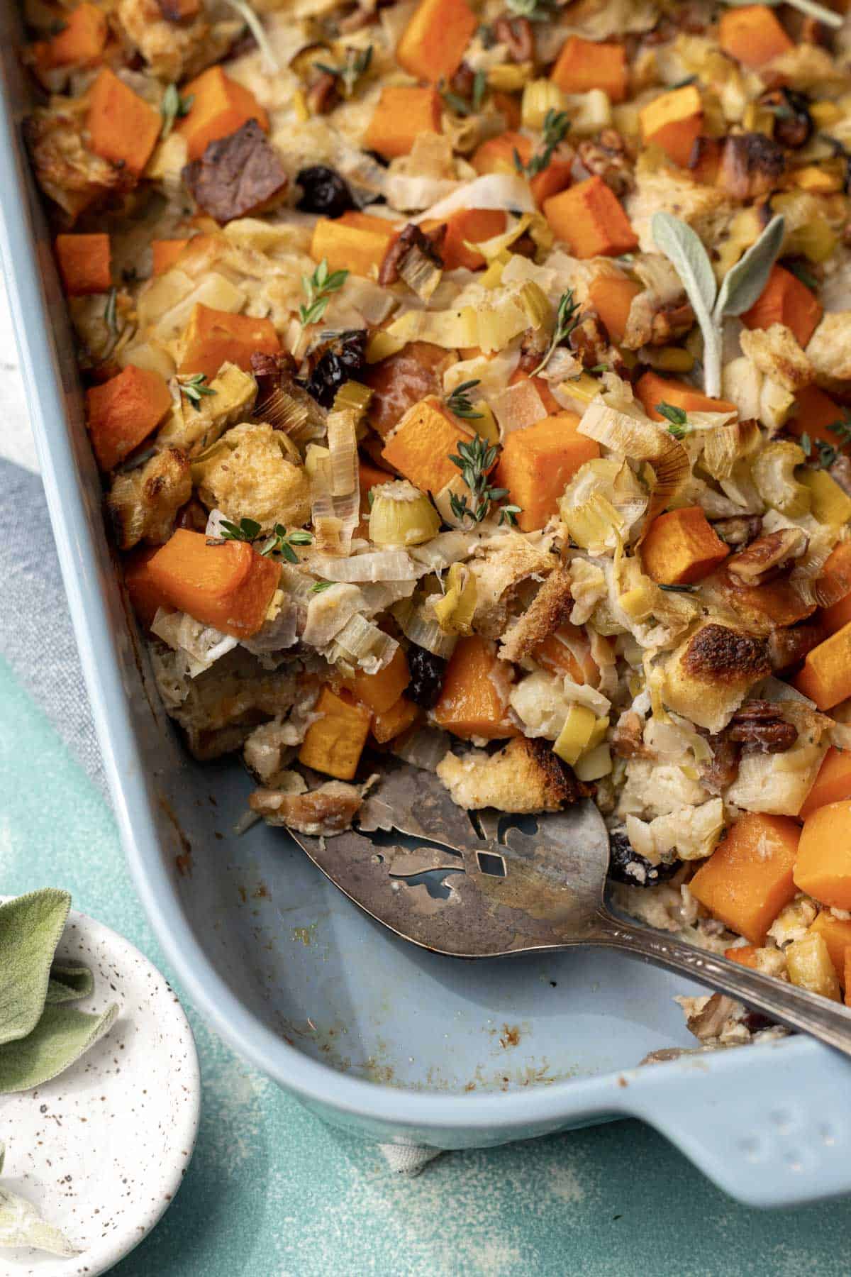 Butternut squash and apple stuffing in a baking dish with a serving spatula and side of fresh sage.