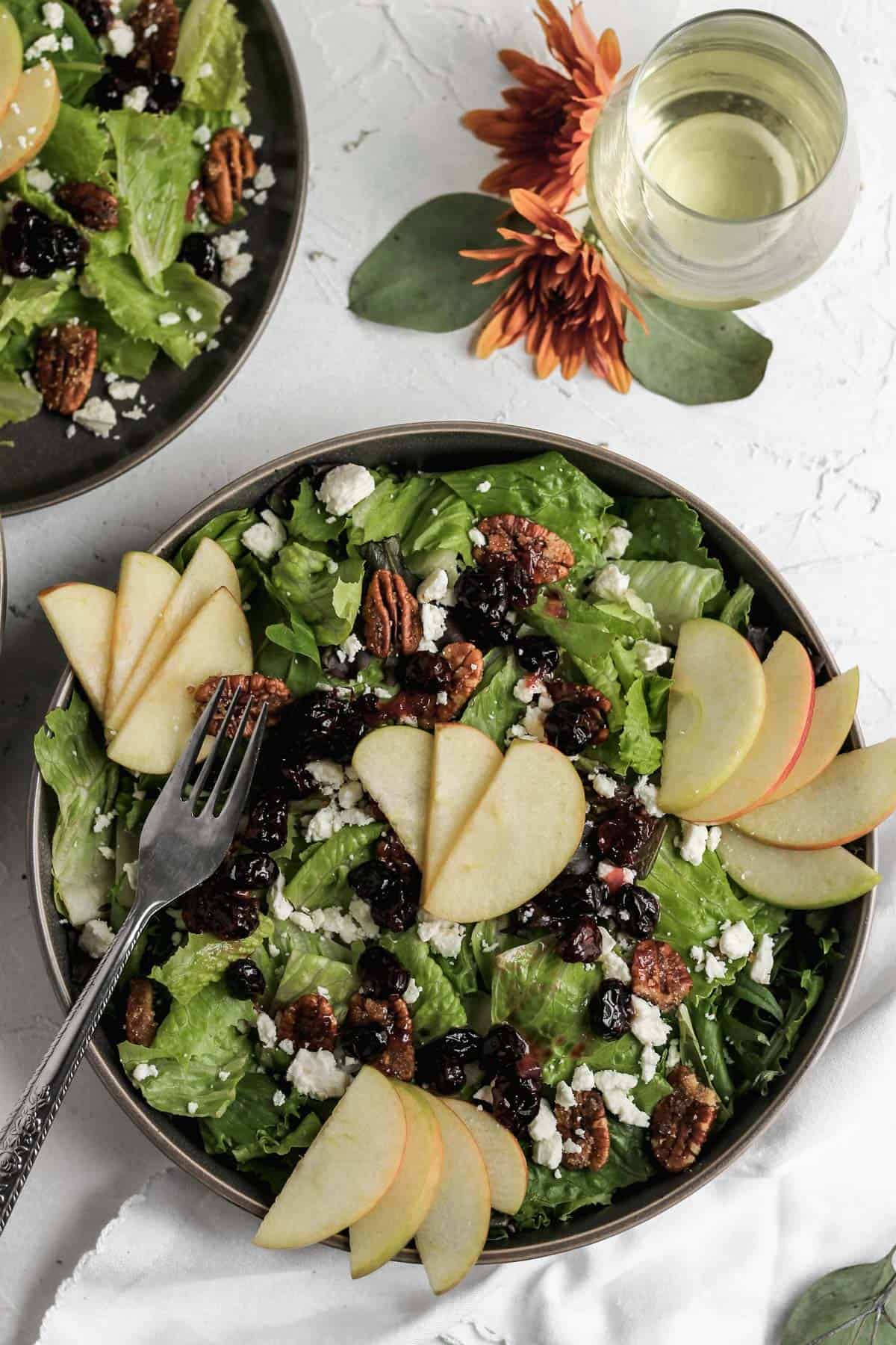 Apple cranberry salad in a black bowl with a silver fork, glass of white wine, and orange flowers.