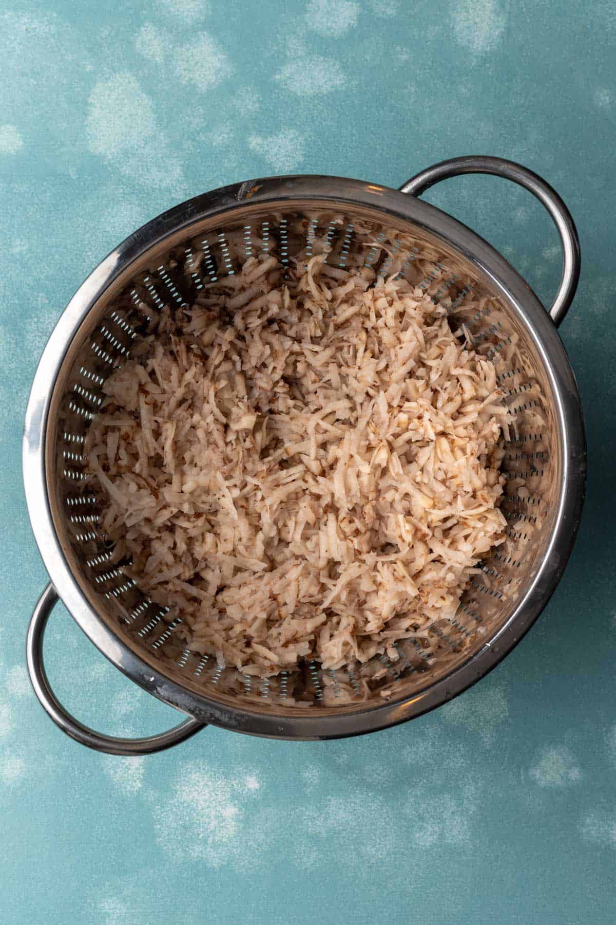 Shredded potatoes and onions in a silver colander.