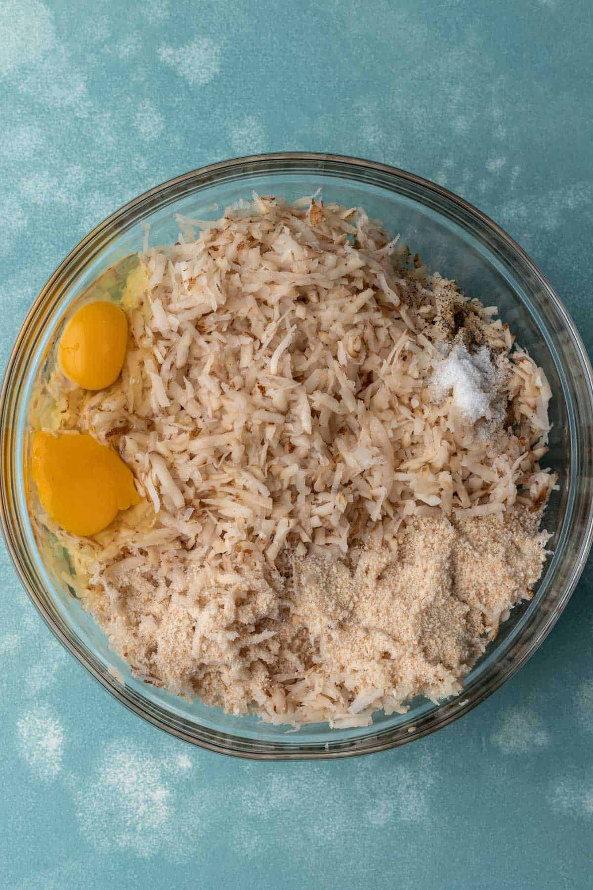 Shredded potatoes and onions with eggs, salt, and breadcrumbs in a large mixing bowl.