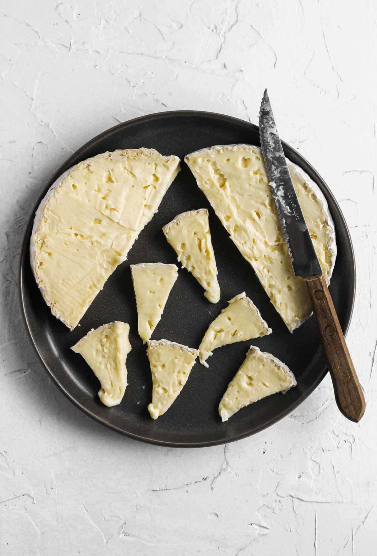 Small triangles of brie cut with a knife on a dark grey plate.