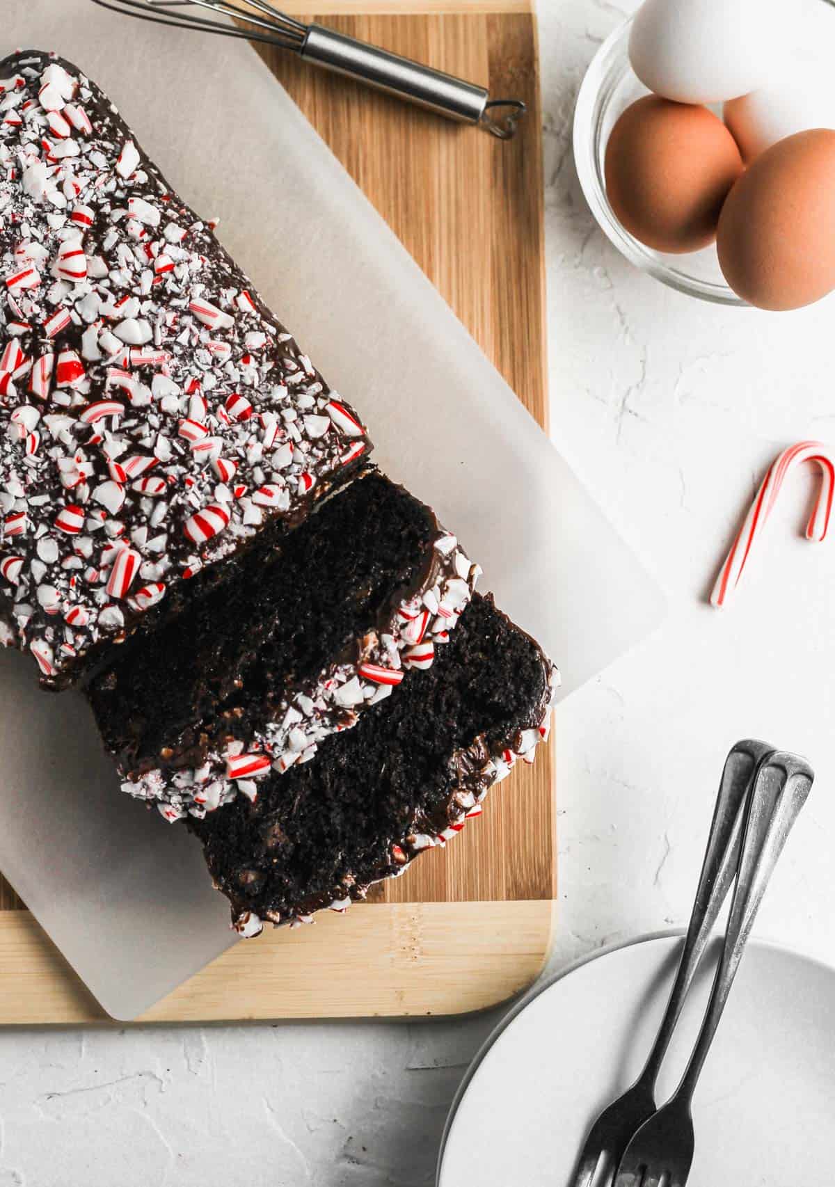 Chocolate zucchini bread with chocolate glaze and crushed peppermints on top with a bowl of eggs and a candy cane next to it.