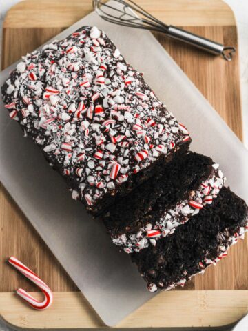 Peppermint death by chocolate zucchini bread on a white cutting board on top of a wooden cutting board with a candy cane next to it.