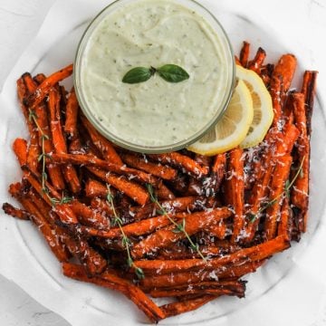 Air fryer carrot fries in a white bowl, garnished with freshly grated parmesan cheese, lemon slices, and a bowl of pesto aioli on the side.