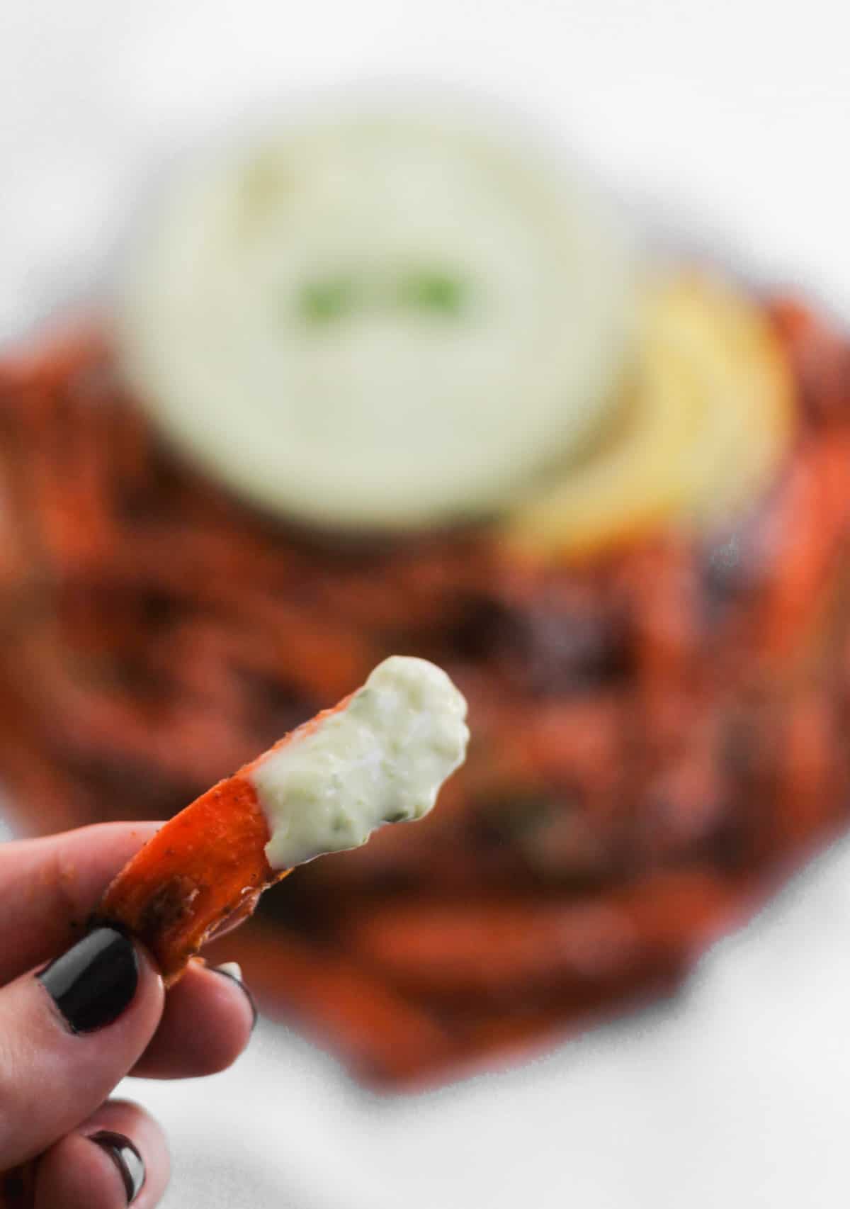 A single carrot fry dipped in pesto aioli with a serving platter of air fryer carrot fries blurred in the background.