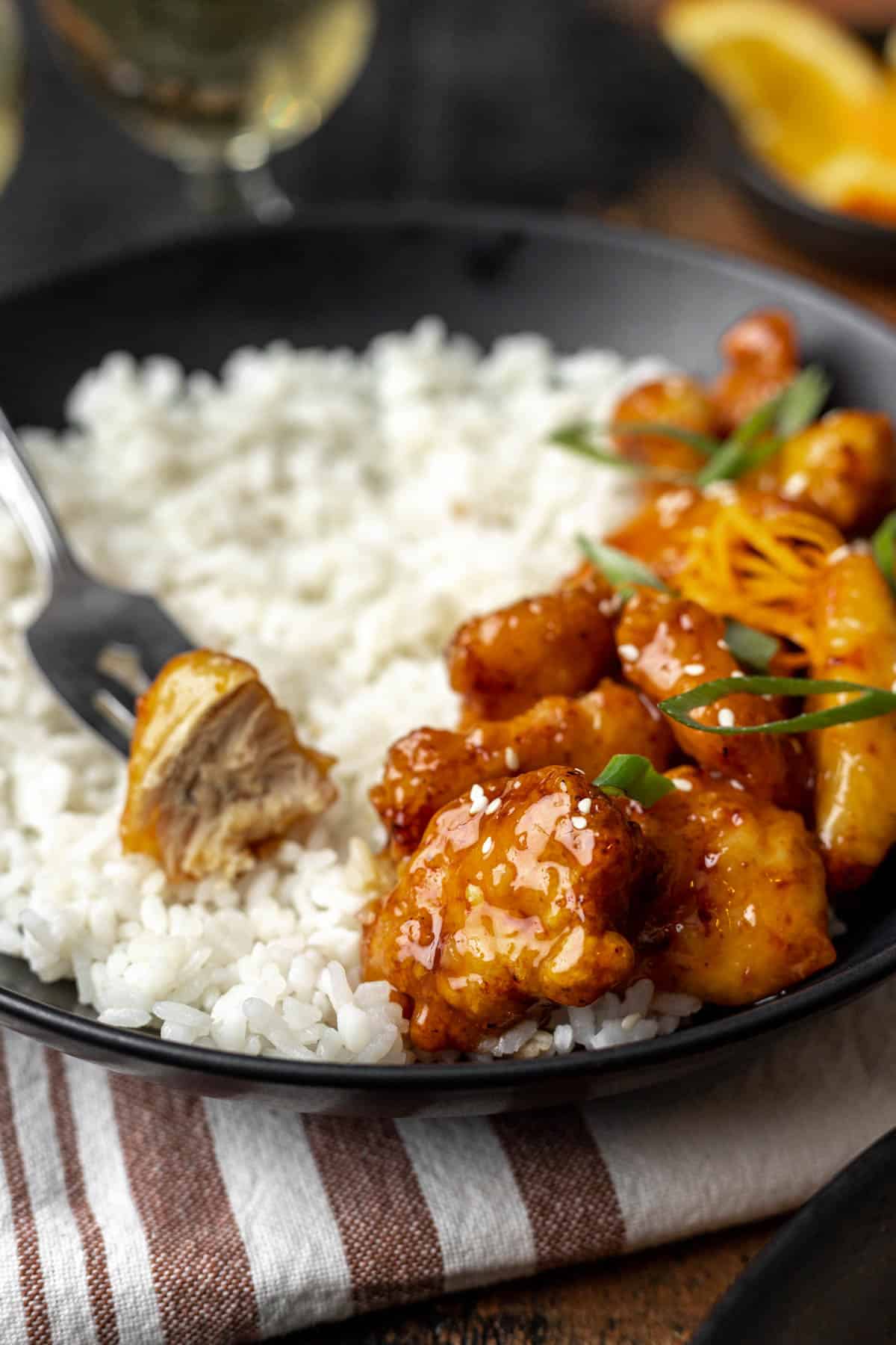 A fork with a piece of orange chicken and a bite taken out next to a pile of more orange chicken.