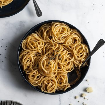 Bucatini cacio e pepe spiraled inside a large blue serving bowl with a serving spoon inside and cheese crumbles around it.