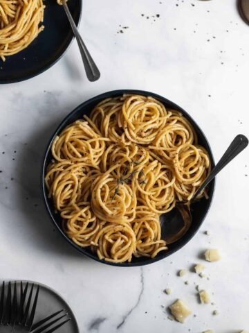 Bucatini cacio e pepe spiraled inside a large blue serving bowl with a serving spoon inside and cheese crumbles around it.