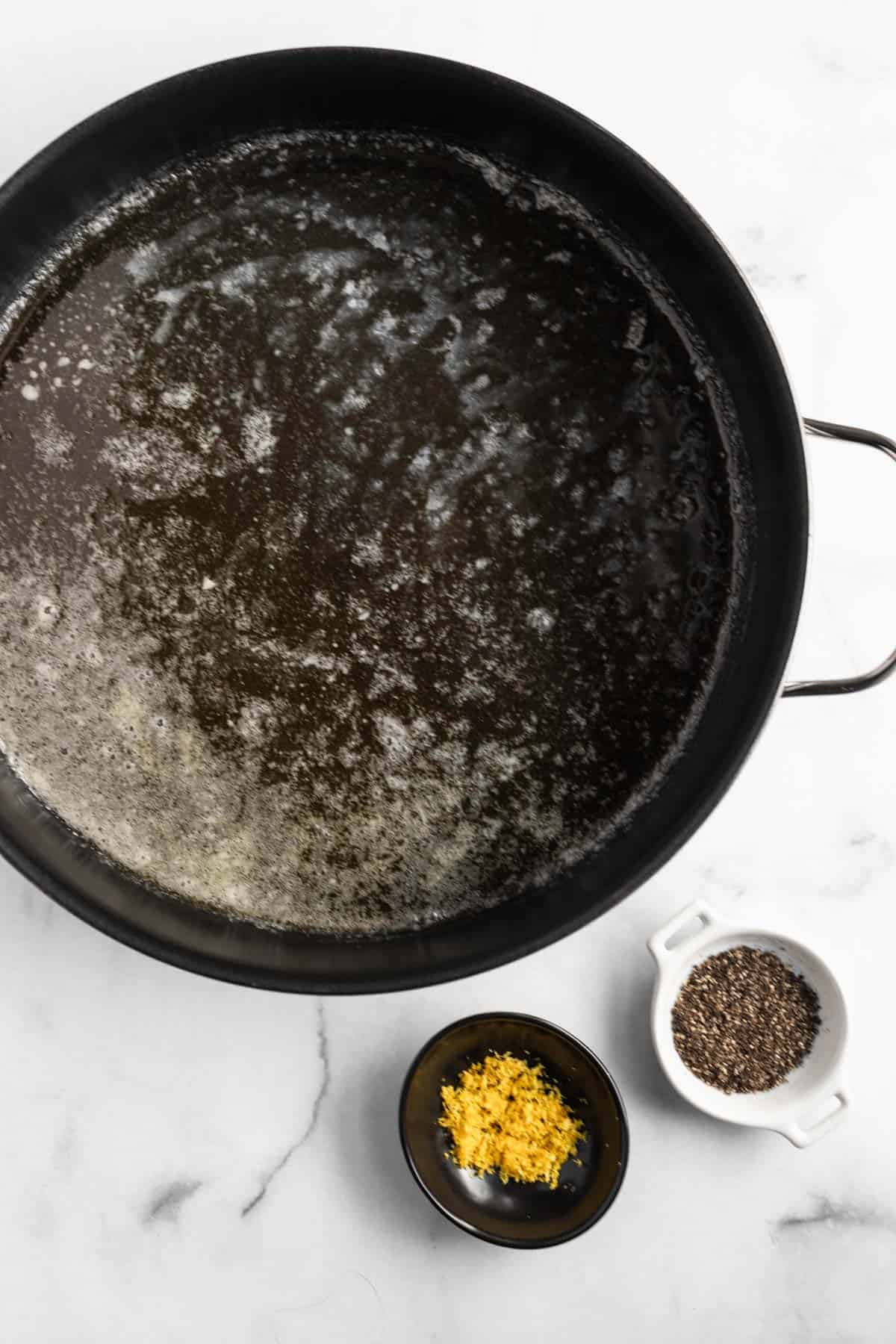 Melted butter in a large black pot with lemon zest and cracked pepper in small bowls off to the side.