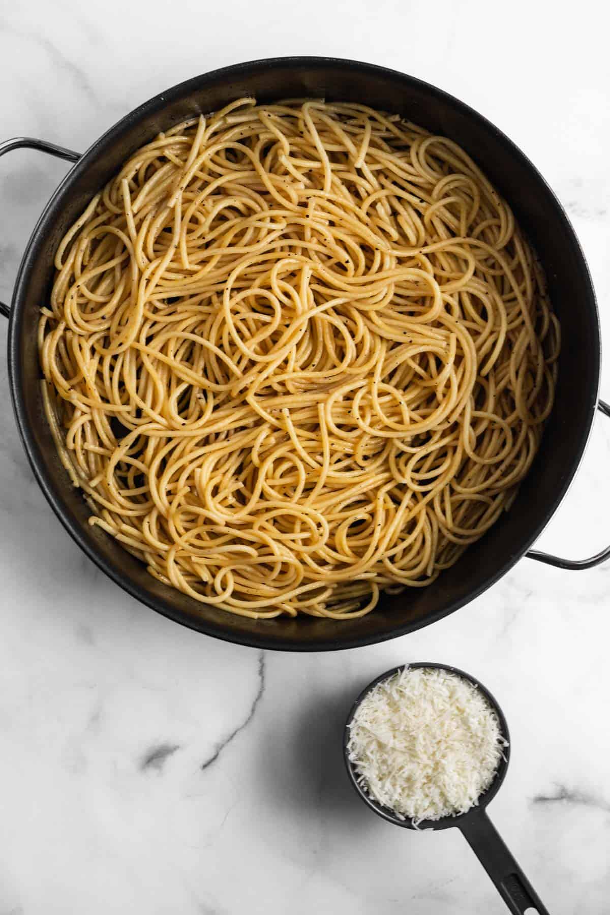 Bucatini noodles added to the melted butter, lemon zest, and cracked pepper in a large black pot with a measuring cup full of freshly grated cheese off to the side.