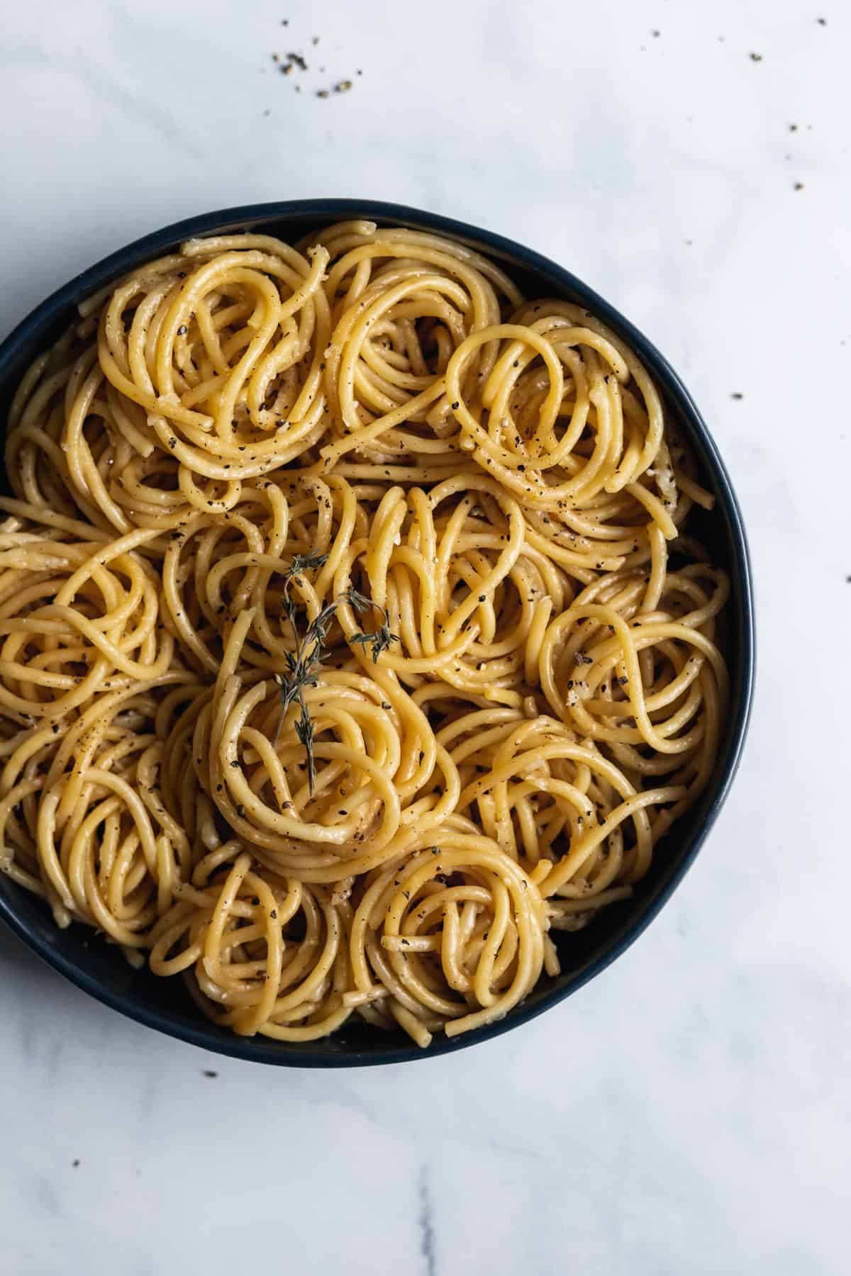 Close up view of a serving bowl filled with spiraled cacio e pepe and garnished with a small sprig of thyme.