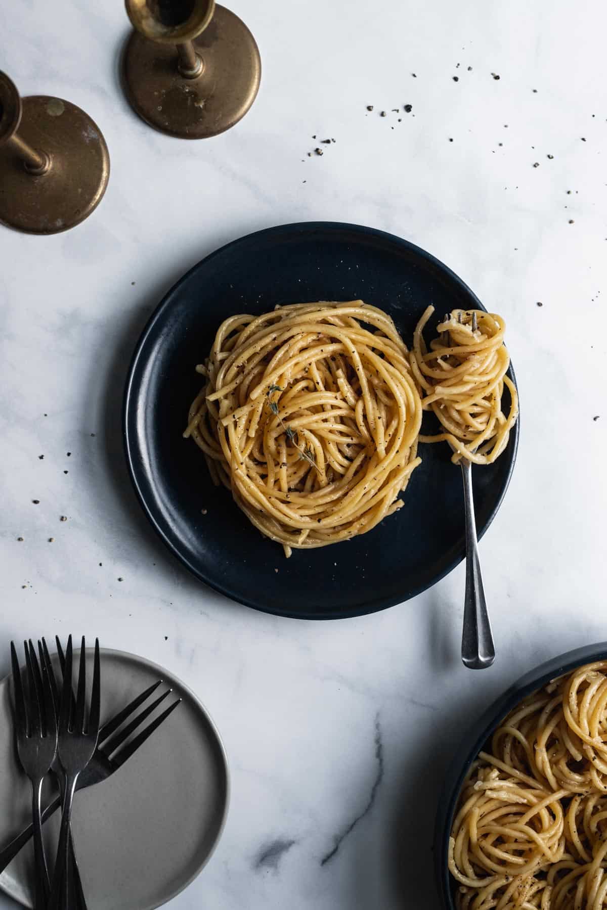 Spiraled bucatini cacio e pepe on a blue plate with a fork twirled in pasta next to it and candle sticks, plates, and a serving dish surrounding it. 