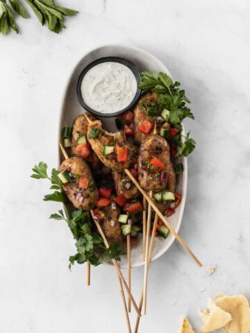 Chicken kofta kebabs on an oval plate garnished with israeli salad and parsley with a side of lemon dill yogurt sauce.