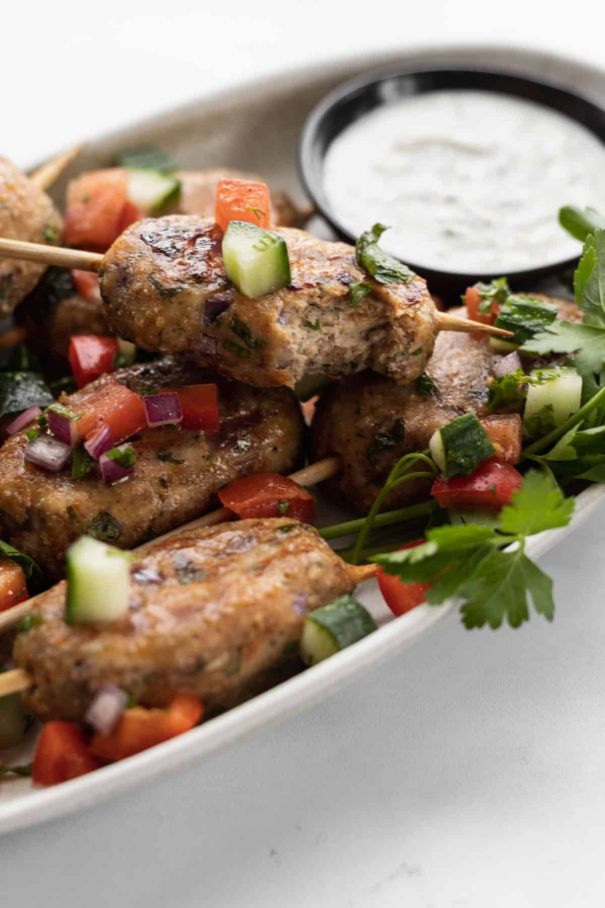 Close up of chicken kofta kebabs on an oval platter garnished with israeli salad and a side of yogurt sauce.