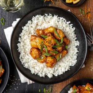 Air fryer orange chicken on a bed of rice in a black bowl topped with scallions.