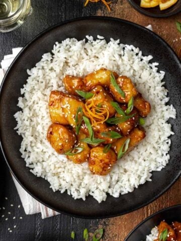 Air fryer orange chicken on a bed of rice in a black bowl topped with scallions.