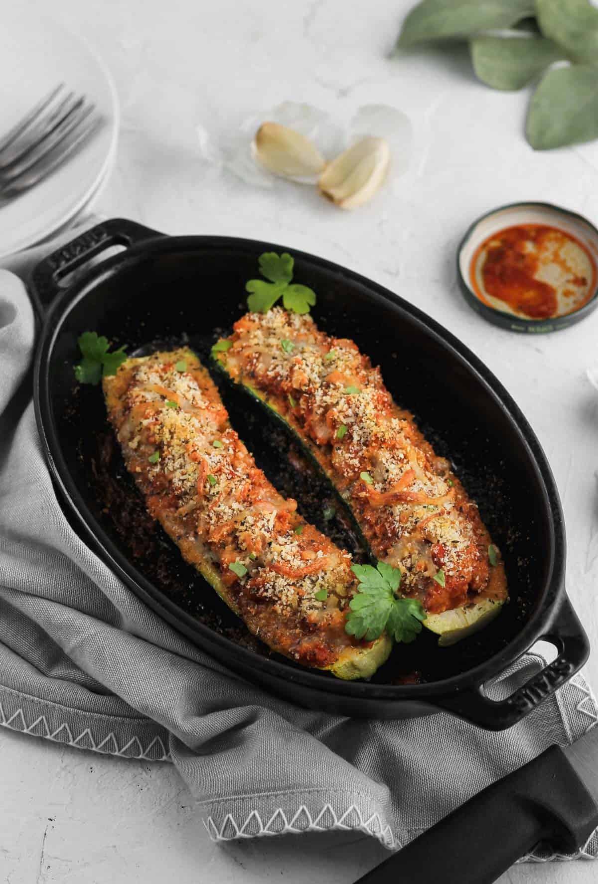 Stuffed zucchini in a black baking dish with a gray napkin and garlic cloves off to the side.