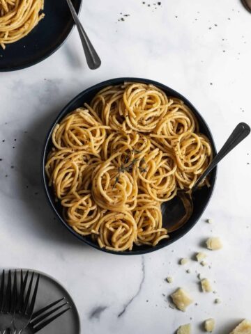 Bucatini cacio e pepe in a blue serving bowl with a spoon and parmesan cheese sprinkled below it.