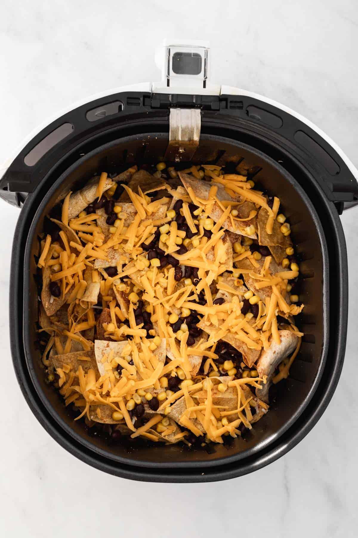 Tortilla chips, black beans, corn, and cheese layered in an air fryer basket.