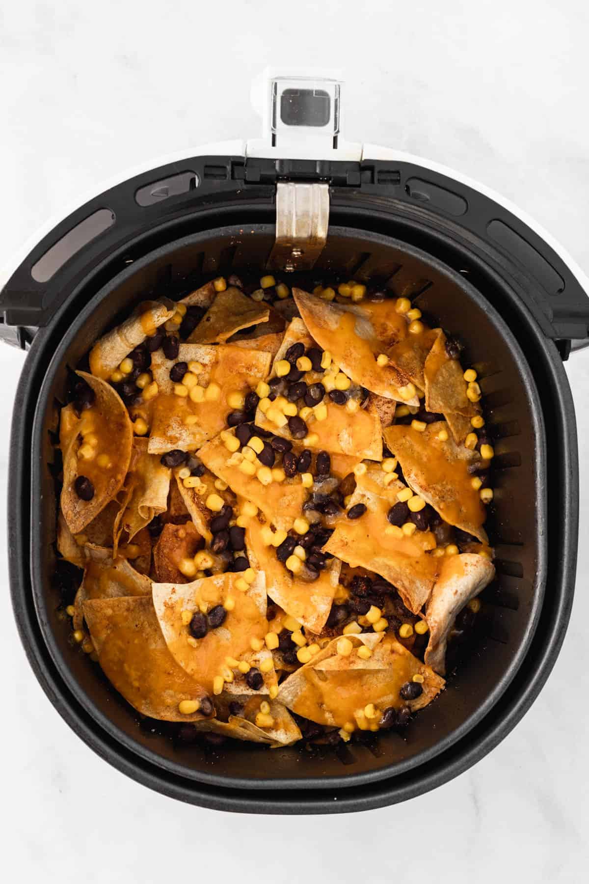 Melted cheese on top of layered tortilla chips, black beans, and corn in an air fryer basket.