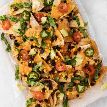 Air fryer nachos with all the fixings on a piece of parchment paper surrounded by a bowl of salsa, sour cream, and Spanish rice.