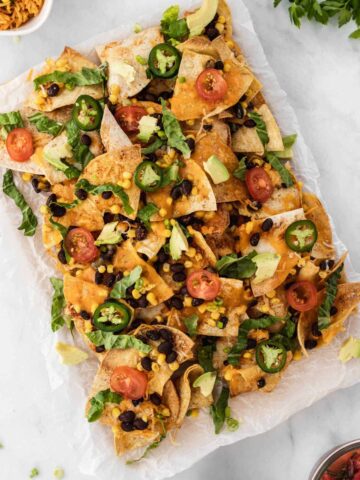 Air fryer nachos with all the fixings on a piece of parchment paper surrounded by a bowl of salsa, sour cream, and Spanish rice.