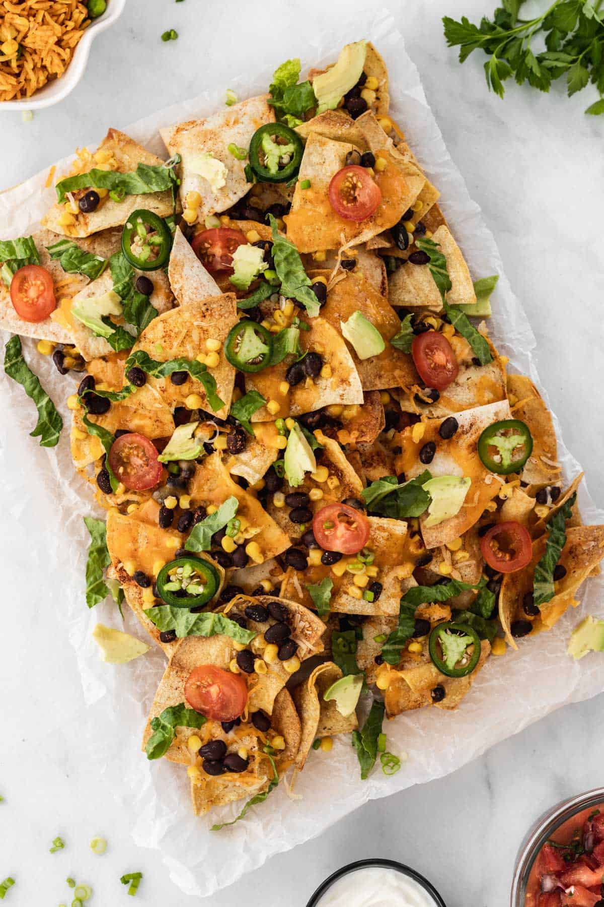Air fryer nachos with all the fixings on a piece of parchment paper surrounded by a bowl of salsa, sour cream, and Spanish rice.