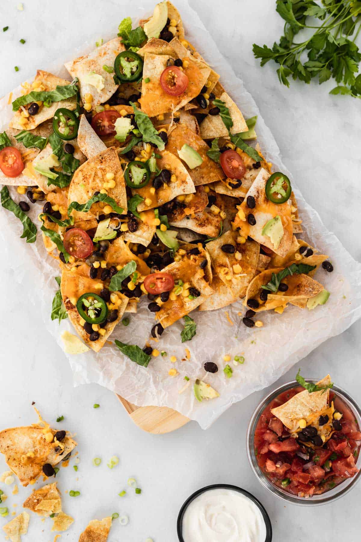 Nachos on top of parchment paper surrounded by broken chips and bowls of sour cream and salsa.
