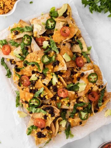 Air fryer nachos with all the fixings on a piece of parchment paper surrounded by a bowl of salsa, sour cream, and Spanish rice.