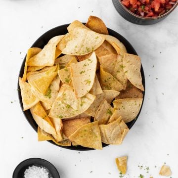 Air fryer tortilla chips in a black bowl with a chip dipped in a bowl of salsa, sea salt, and limes around it.