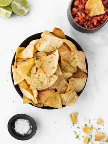 Air fryer tortilla chips in a black bowl with a chip dipped in a bowl of salsa, sea salt, and limes around it.