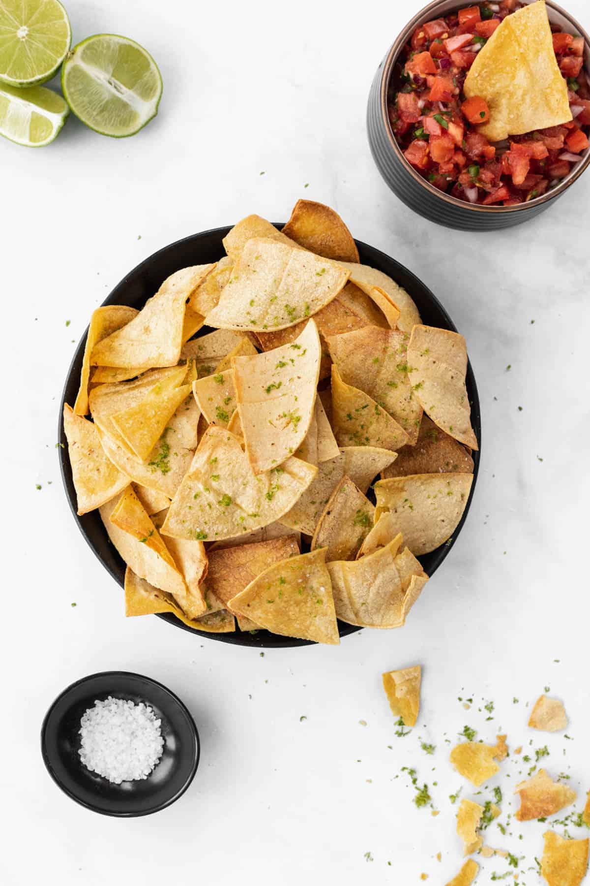 Air fryer tortilla chips in a black bowl with sea salt, limes, and a chip dipped in a bowl of salsa around it.