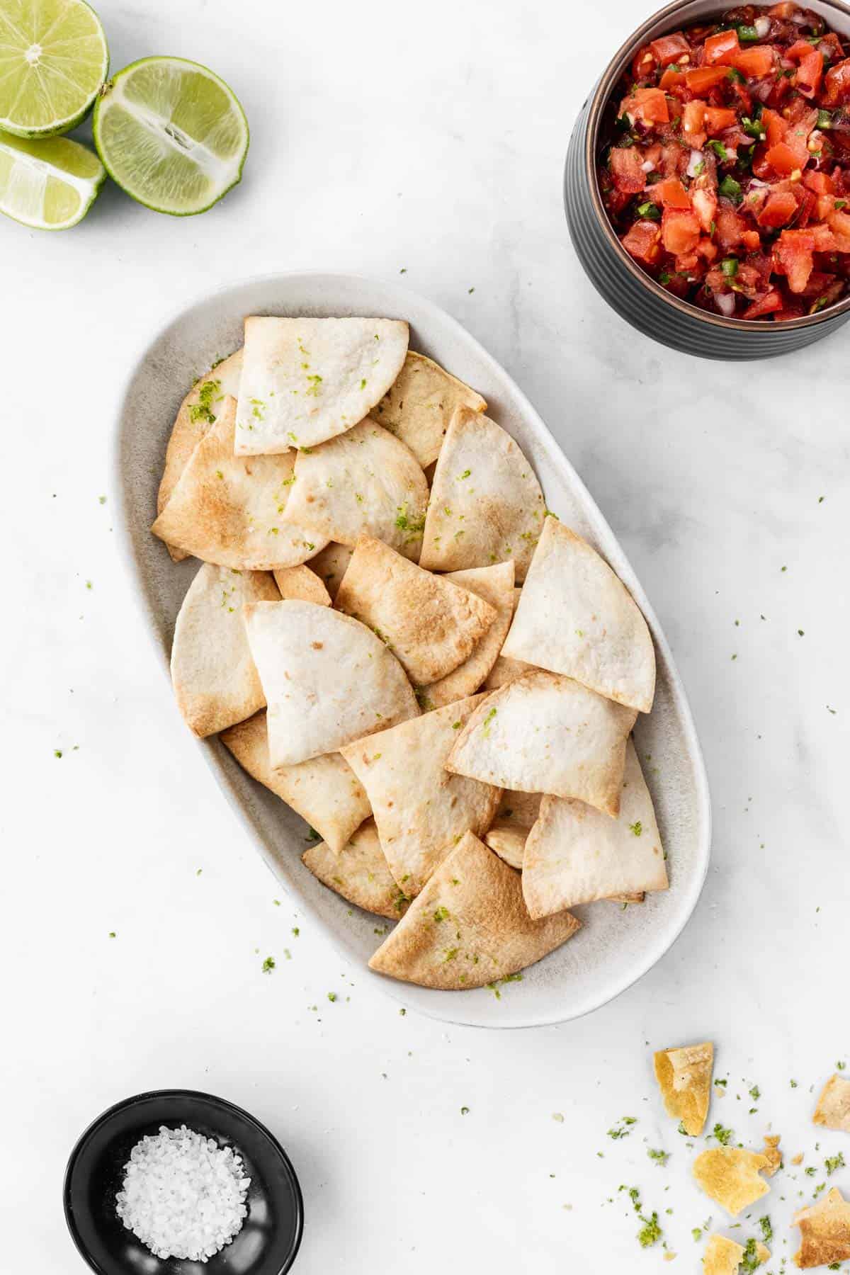 Air fryer flour tortilla chips on an oval platter with sea salt, limes, and a bowl of salsa around it.