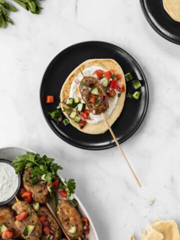 Chicken kofta kebab on a blue plate with a small pita, lemon yogurt sauce, and israeli salad on top.