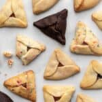 Different kinds of hamantaschen scattered on a white surface with some crumbs.