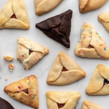 Different kinds of hamantaschen scattered on a white surface with some crumbs.