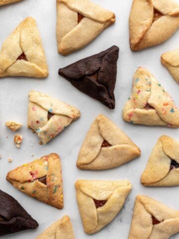 Different kinds of hamantaschen scattered on a white surface with some crumbs.
