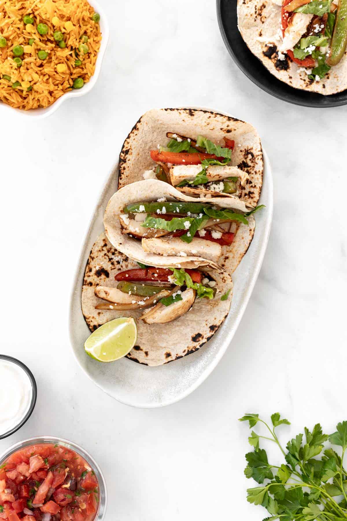 3 fajita tacos on a serving platter surrounded by another taco on a separate plate, a bunch of cilantro, and bowls of salsa, sour cream, and spanish rice.