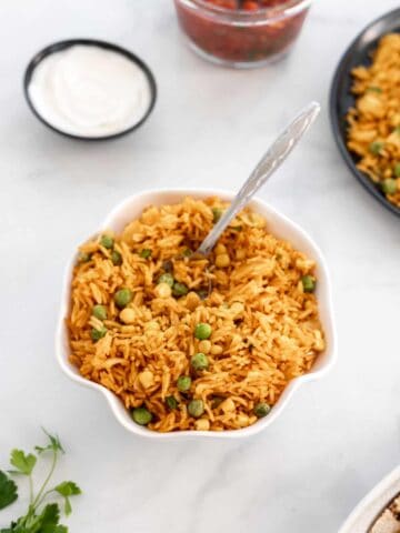 Instant pot Spanish rice in a white ruffled bowl surrounded by cilantro and small bowl of sour cream.