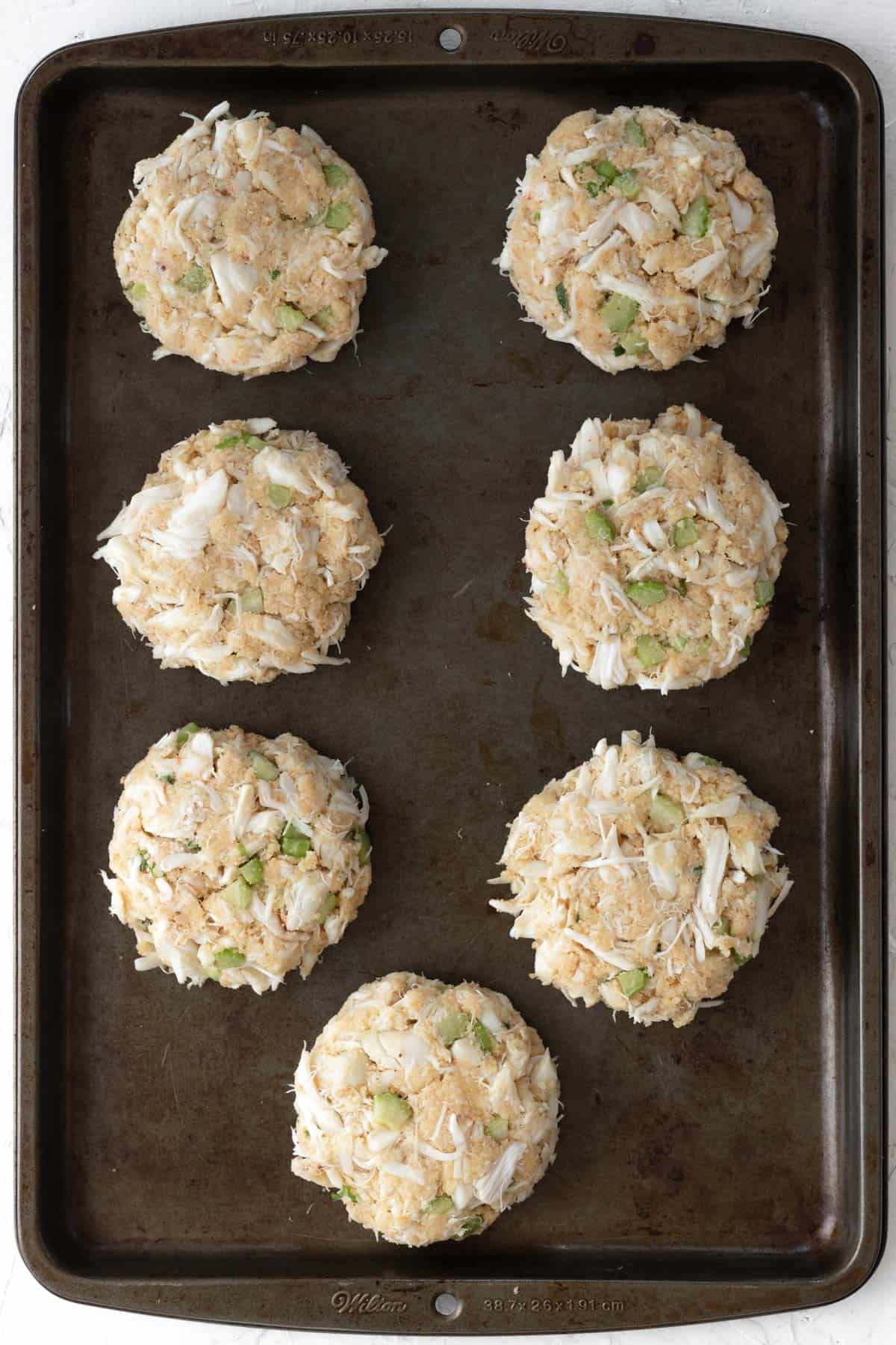 7 raw crab cakes on a metal baking sheet.