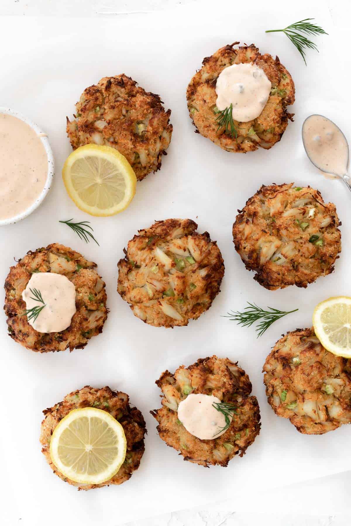 Air fryer crab cakes on a white backdrop with a bowl of remoulade sauce, lemon slices, and dill sprigs around them.